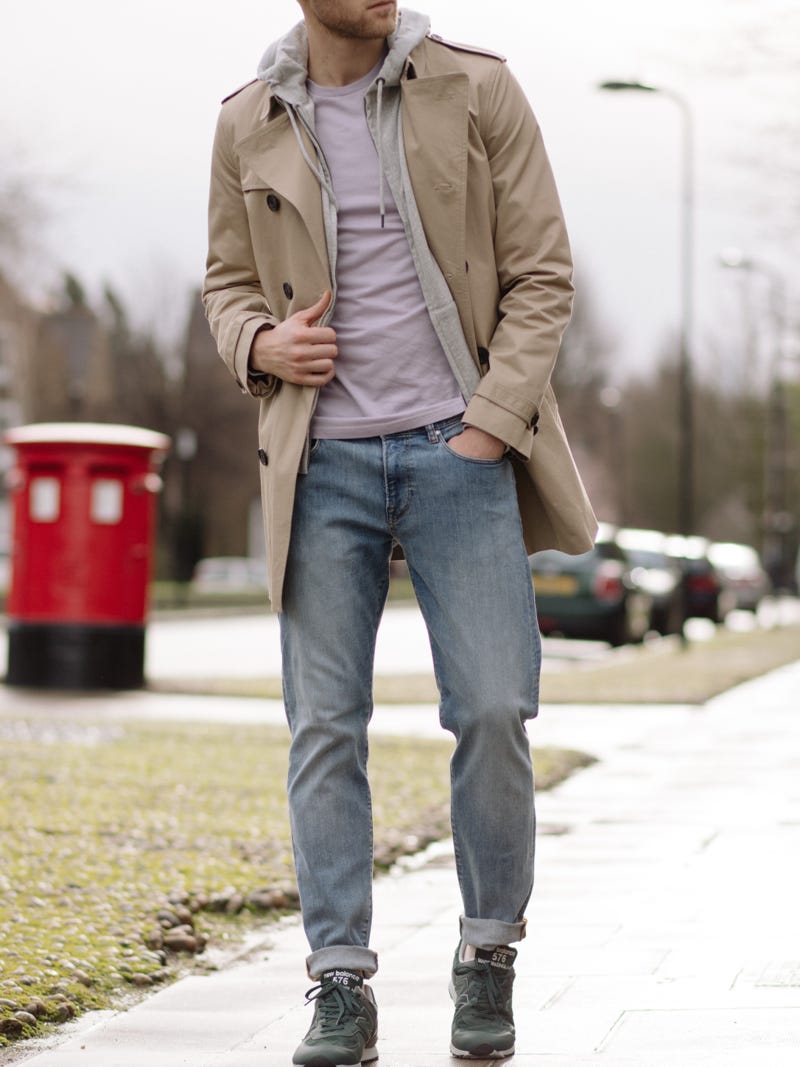 Men's outfit idea for 2022 with stone trench coat, gray hoodie, pale-colored crew neck t-shirt, light blue jeans, green everyday sneakers. Suitable for spring and fall.